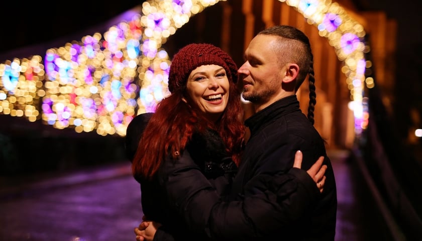 Radosna para w objęciach: rudowłosa kobieta  i mężczyzna z warkoczem, w tle podświetlony na różne kolory most Tumski