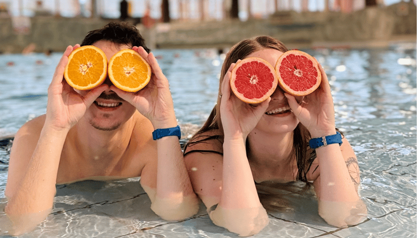 Para na basenie, leżąca w wodzie i trzymająca połówki owoców jak okulary