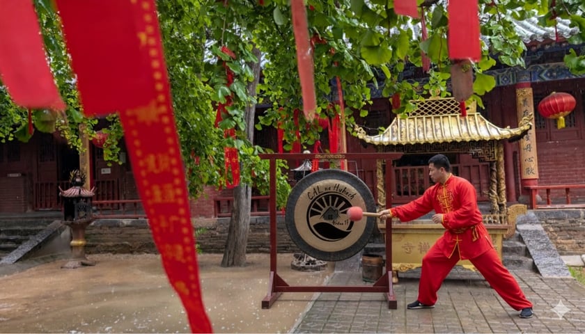Na dziedzińcu w stylu chińskim, wśród zielonych drzew i czerwonych wstęg z chińskimi znakami, stoi duży gong z logo „CAL na Hubach Widna 17”. Mężczyzna w czerwonym stroju do sztuk walki, uderza w gong pałką.