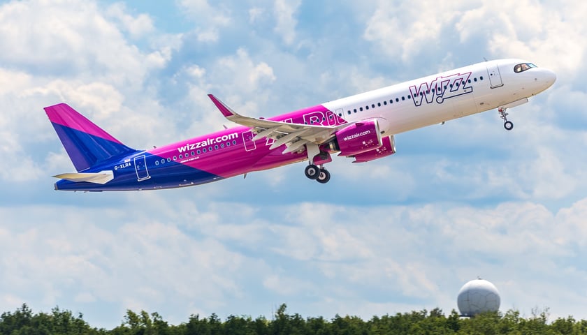 Startujący samolot pomalowany na biało, fioletowo i niebiesko węgierskiej taniej linii lotniczej Wizzair