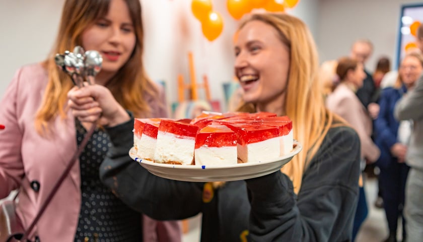 Dwie młode kobiety, jedna stoi z tyłu, duga trzyma łyżeczki i niesie talerz z ciastem.