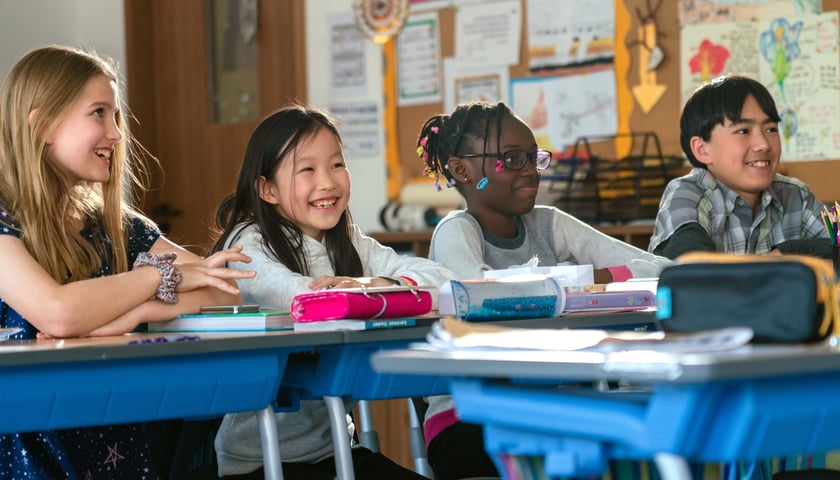 Zdjęcie ilustracyjne - czwórka uśmiechniętych dzieci różnych ras siedząca w szkolnych ławkach na lekcji.