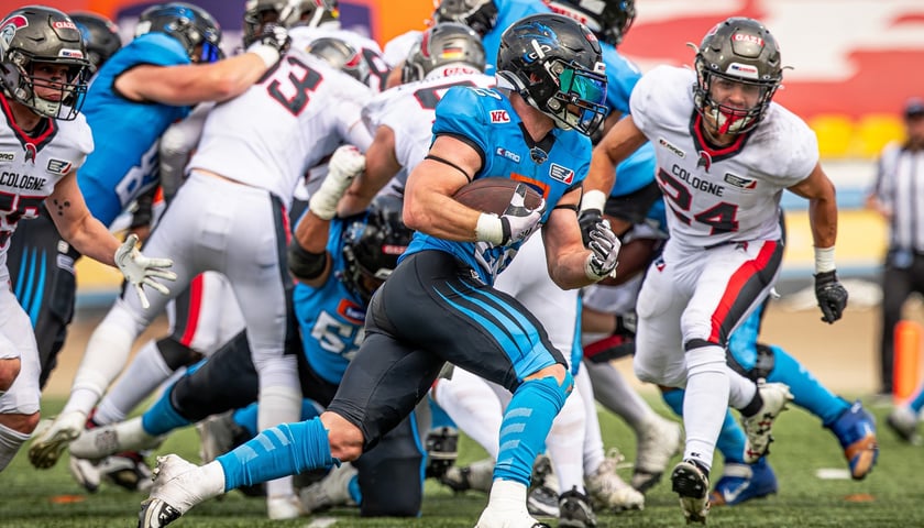 Zawodnik Panthers Wrocław biegnie z piłką do gry w futbol amerykański w meczu z Cologne Centurions w lidze ELF. W tle walka zawodników obu drużyn.