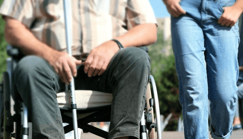 Na zdjęciu mężczyzna na wózku inwalidzkim z laską. Zdjęcie ilustracyjne