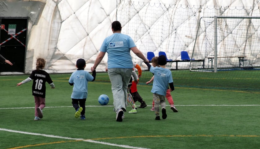 Zdjęcie ilustracyjne - grupa dzieci biegnących z osoba dorosłą po boisku piłkarskim - poprzedni turniej Piłka Bez Barier.