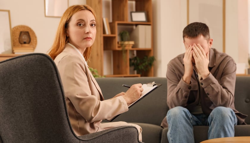 Psycholog kobieta w beżowym garniturze siedzi z notatnikiem i patrzy w stronę aparatu fotograficznego, podczas gdy mężczyzna w brązowej marynarce siedzi obok niej na kanapie z rękami zasłaniającymi twarz, wyrażając smutek lub stres.