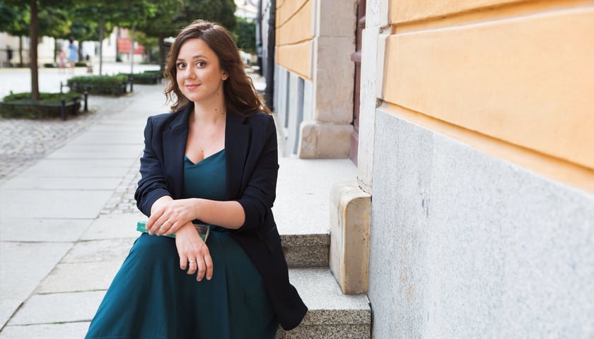 Maria Paszyńska. Na zdjęciu młoda kobieta w sukience siedzi na schodku przed wejściem do budynku