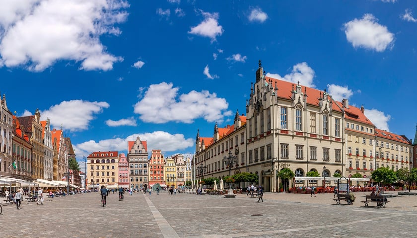Panoramiczny widok Rynku we Wrocławiu w słoneczny dzień. Na pierwszym planie brukowany plac z przechodniami i rowerzystami. Po lewej stronie kolorowe kamienice mieszczańskie z czerwonymi dachami i kawiarniami pod parasolami. Po prawej stronie Ratusz Wrocławski z charakterystyczną wieżą zegarową i zielonymi iglicami.