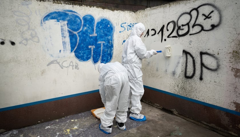 Dwie osoby w białych kombinezonach i maskach zamalowują napisy na murach