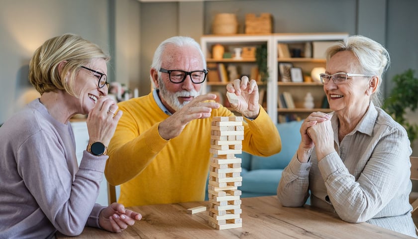 Dwie uśmiechnięte kobiety w okularach i mężczyzna przy stole układają drewniane klocki