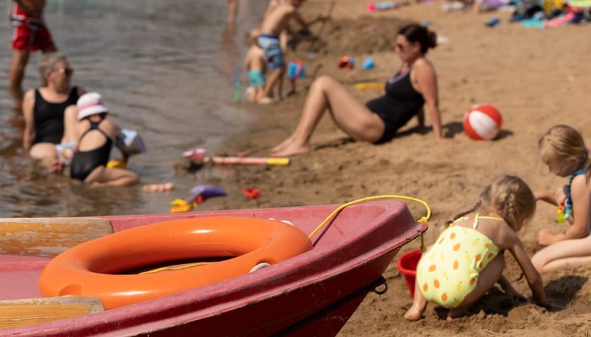 Powiększ obraz: Archiwalne zdjęcie przedstawia wypoczywających na plaży kąpieliska Glinianki ludzi, z widoczną na pierwszym planie łodzią ratunkową i bawiącymi się w piasku dziećmi