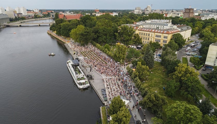 Powiększ obraz: Zdjęcie z lotu ptaka przedstawia koncert na statku zacumowanym przy bulwarze; na schodach i nadbrzeżu zgromadził się tłum ludzi