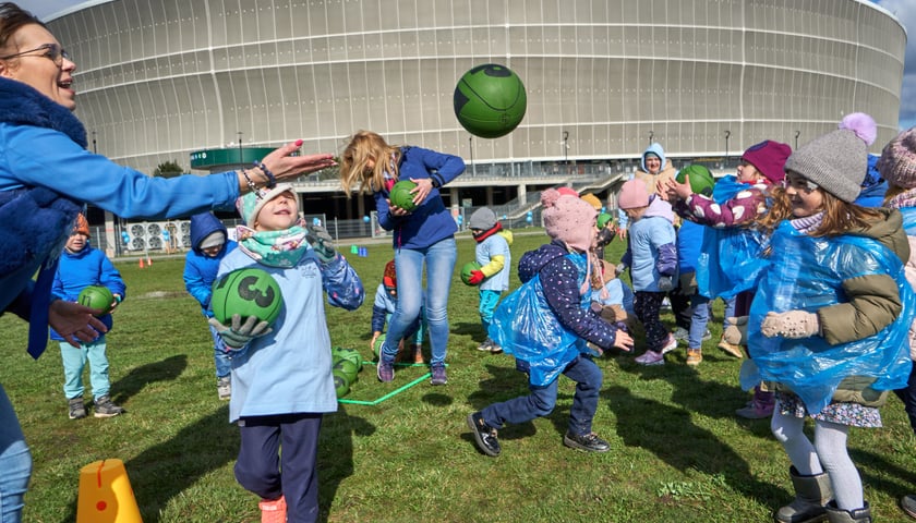 Powiększ obraz: Na zdjęciu przedszkolaki w niebieskich foliowych płaszczach grają zieloną piłką na trawniku przed Tarczyński Arena
