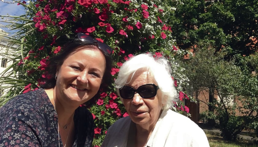 Powiększ obraz: Na zdjęciu dwie kobiety: wolontariuszka Aneta i seniorka Irena na tle kwitnącego na różowo krzewu