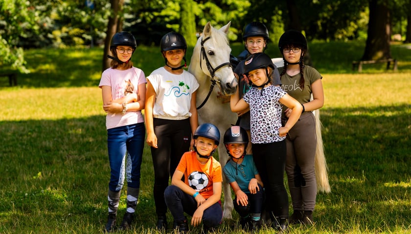 Powiększ obraz: Na zdjęciu siedmioro dzieci  wokół siwego konia na trawie w cieniu drzewa