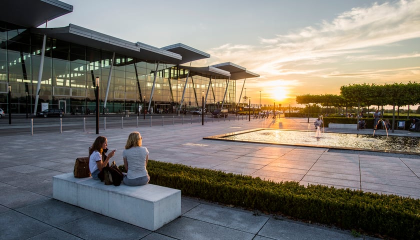 Powiększ obraz: Ludzie siedzący przed wrocławskim lotniskiem o zachodzie słońca