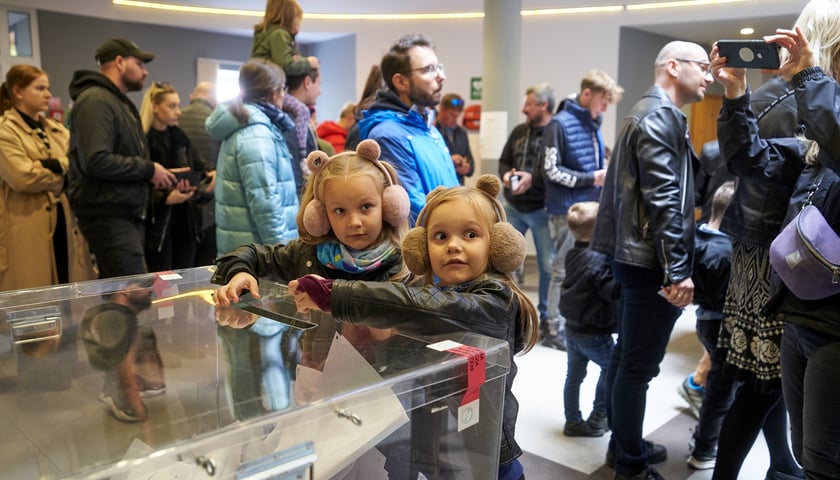 Powiększ obraz: Na zdjęciu dwie małe dziewczynki przy urnie wyborczej, w tle kolejka do głosowania w lokalu wyborczym przy ul. Gazowej
