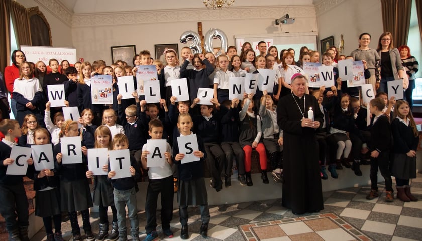 Dzieci trzymające karteczki z literami mającymi układać się w napis związany ze świecami wigilijnymi Caritas
