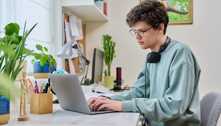 Powiększ obraz: Student w okularach i ze słuchawkami na szyi przy otwartym laptopie