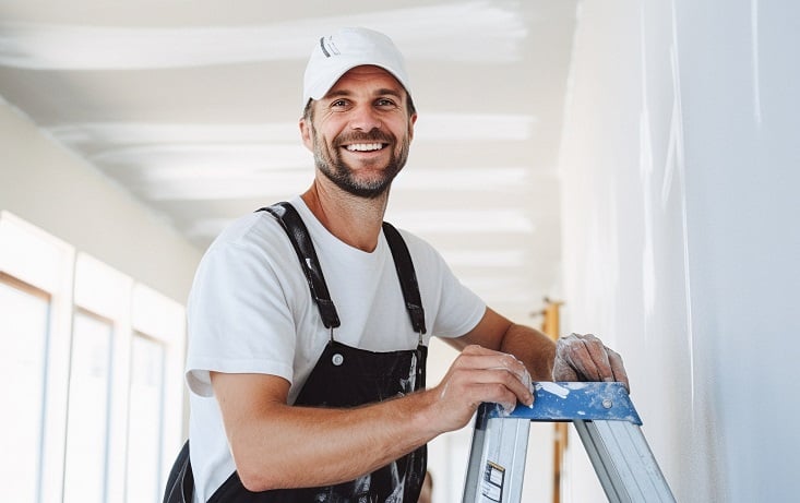 Powiększ obraz: Uśmiechnięty mężczyzna w stroju roboczym umazanym białą farbą, stojący na drabinie