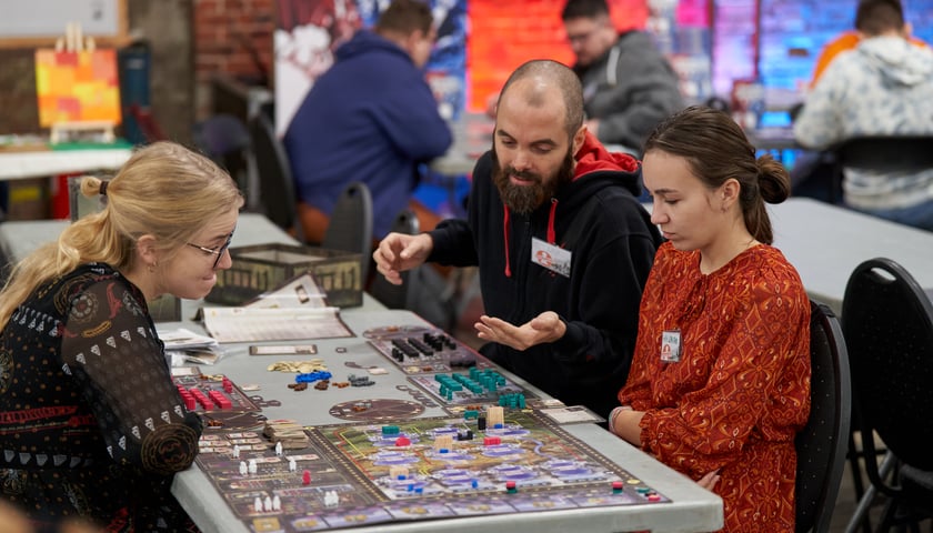 Powiększ obraz: Trzy osoby przy stoliku z planszówkami - Wrocław Games Fest 2023