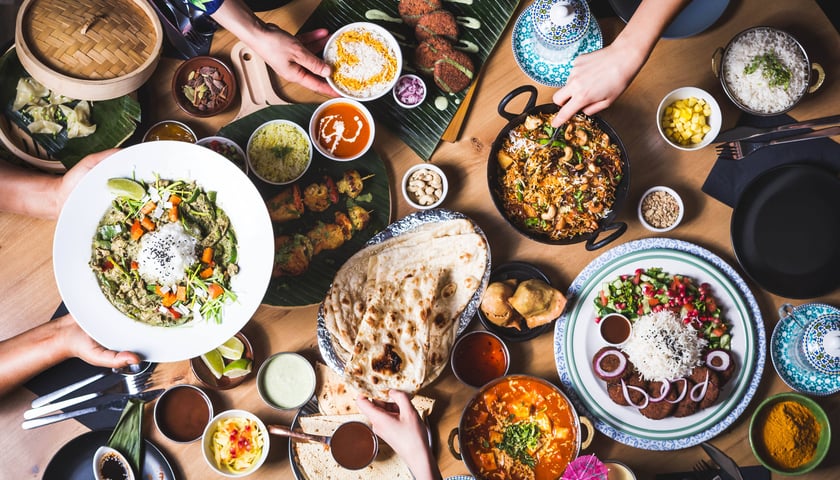 Powiększ obraz: Zdjęcie przedstawia stół nakryty półmiskami pełnymi dań kuchni indyjskiej z wrocławskiej restauracji Mango Mama.