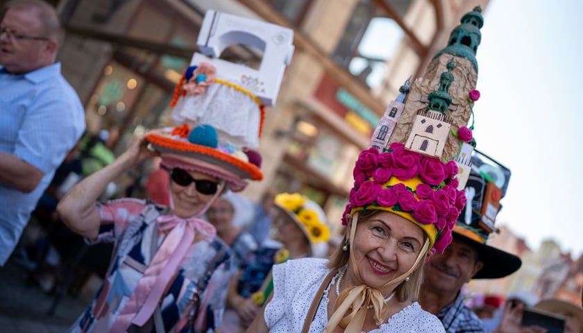 Powiększ obraz: Dwie kobiety w średnim wieku w fantazyjnych kapeluszach (jedna ma go w kształcie budynku kościoła w kwiatach)