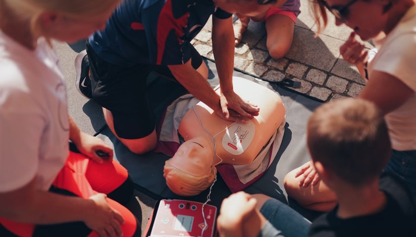 Powiększ obraz: Mężczyzna z ułożonymi dłońmi na fantomie pokazuje dzieciom, jak prowadzić resuscytację