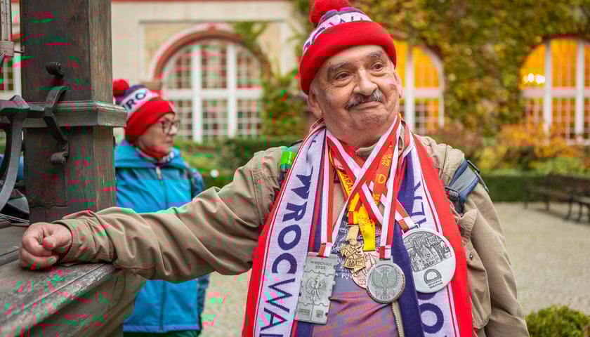 Powiększ obraz: Mężczyzna z wąsem w czapce i szaliku z napisem WrocWalk z 5 medalami WrocWalk zdobytymi we wcześniejszych edycjach