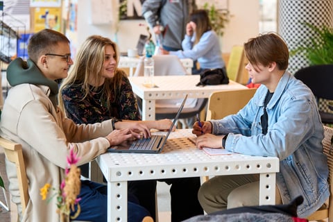 Powiększ obraz: Troje studentów: Jan, Marta i Kacper siedzą przy stoliku w kawiarni; Jan patrzy w ekran laptopa, Marta rozmawia z Kacprem
