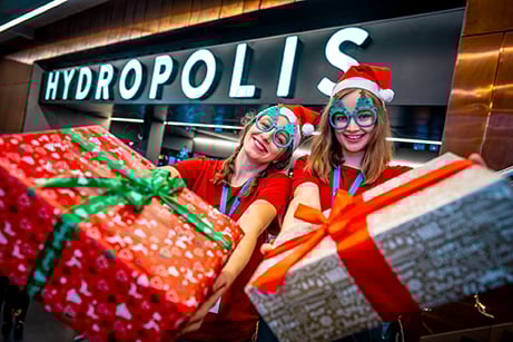 Powiększ obraz: Dwie dziewczyny w czapkach mikołaja i w okularach z choinkami trzymają wielkie pudła prezentowe opakowane kolorowym papierem; w tle napis "Hydropolis"