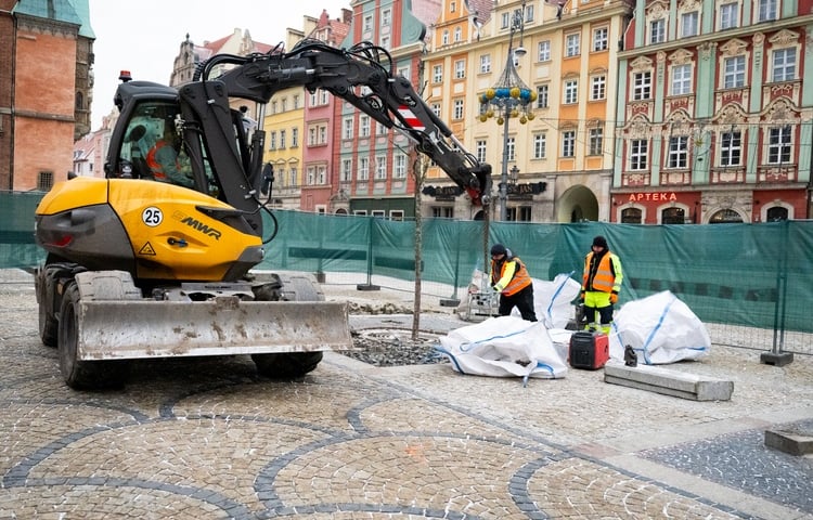 Powiększ obraz: Dwóch robotników w jaskrawych kamizelkach  pracujących przy  wyciąganiu kostki brukowej z płyty Rynku, obok dźwig