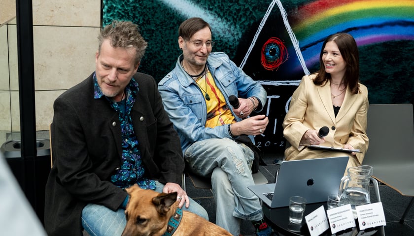 Powiększ obraz: Trzy osoby (organizatorzy) 46. Przeglądu Piosenki Aktorskiej siedzą na tle kolorowej wizualnej kreacji tegorocznej edycji wydarzenia.