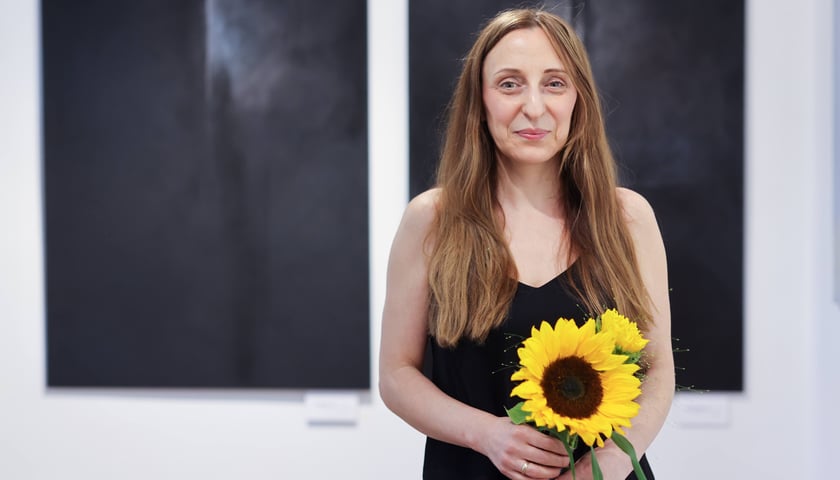 Agnieszka Semaniszyn-Konat ze słonecznikiem na tle swoich obrazów w Galerii Tkackiej na Jatkach, lipiec 2026