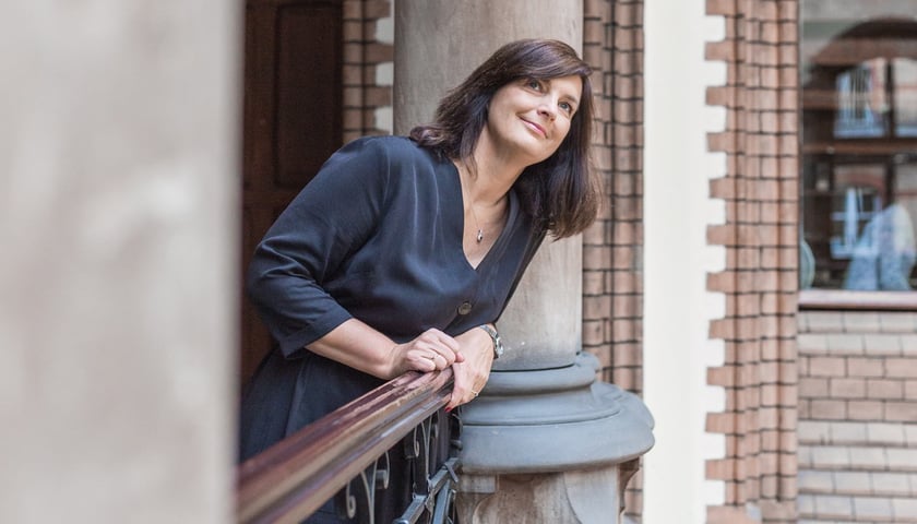 Uśmiechnięta Joanna Kuciel-Frydryszak ubrana w czarną sukienkę wychyla się przez balustradę w dawnej Bibliotece Uniwersyteckiej