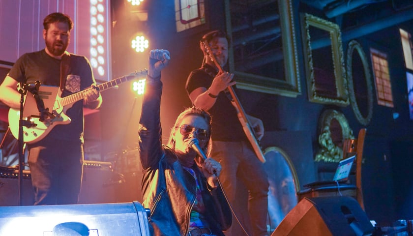 Wokalistka w okularach słonecznych, unosi pięść, za nią dwójka gitarzystów. Wszyscy stoją na scenie podczas koncertu