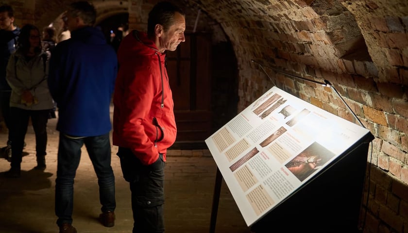 Noc Muzeów 2025 - krypty Dominikanów. Na zdjęciu mężczyzna w czerwonej kurtce w pogrążonych w półmroku podziemiach 