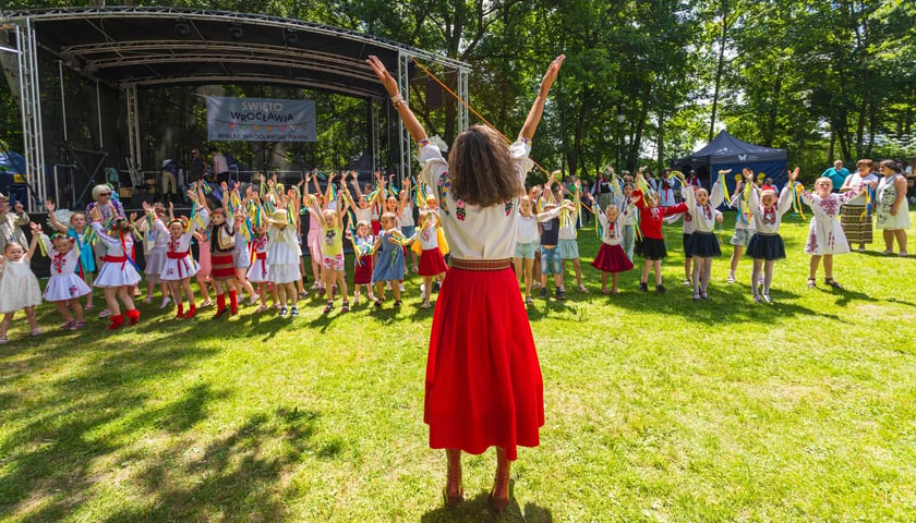 Przed grupą tańczących dzieci stoi kobieta w ludowym stroju i wyciąga ręce do góry