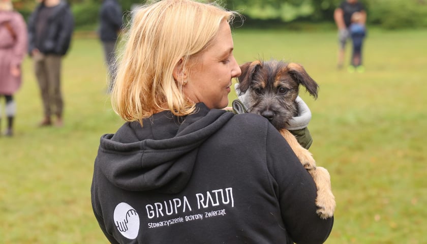 Młoda blondynka stoi plecami do zdjęcia. Ma na sobie bluzę z napisem GRUPA RATUJ i trzyma na rękach małego pieska.