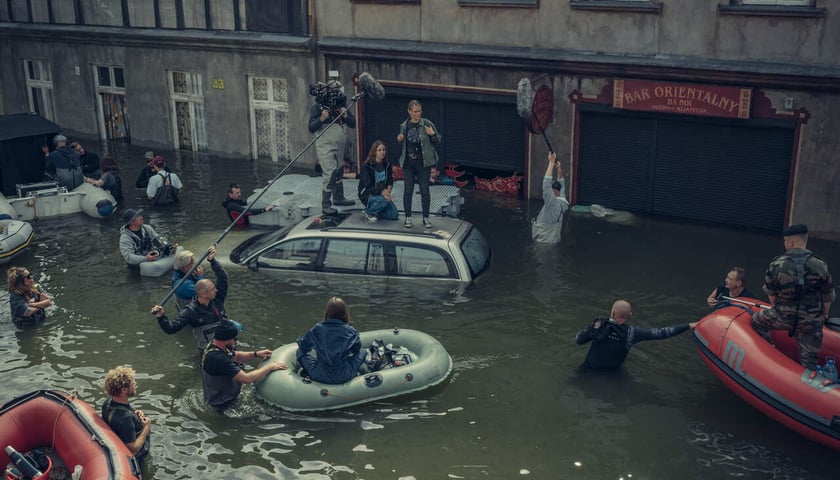 Aktorki Agnieszka Żulewska i Blanka Kot stoją na samochodzie zatopionym przez powódź, wokół pracuje ekipa filmowa. Kręcono w kąpielisku w Srebrnej Górze, gdzie odtworzono Przedmieście Oławskie 