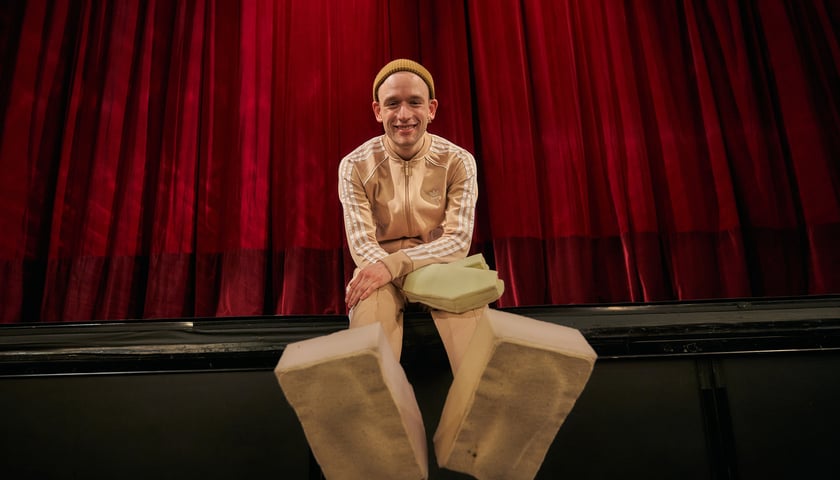 Na zdjęciu Krzysztof Kozak w roli Pinokia siedzi na scenie w charakterystycznych butach z drewna