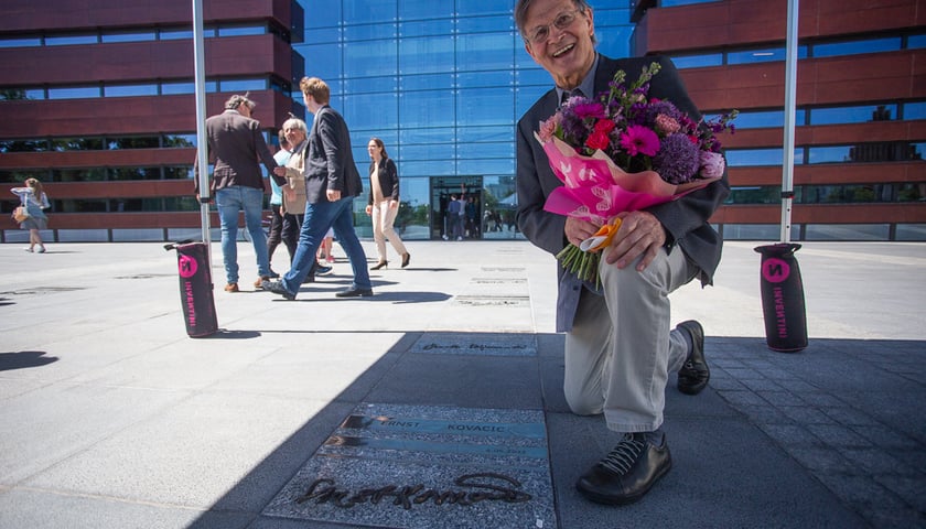 Uśmiechnięty Ernst Kovacic z bukietem kwiatów w ręku przyklęka przy własnej tablicy honorowej przed budynkiem Narodowego Forum Muzyki