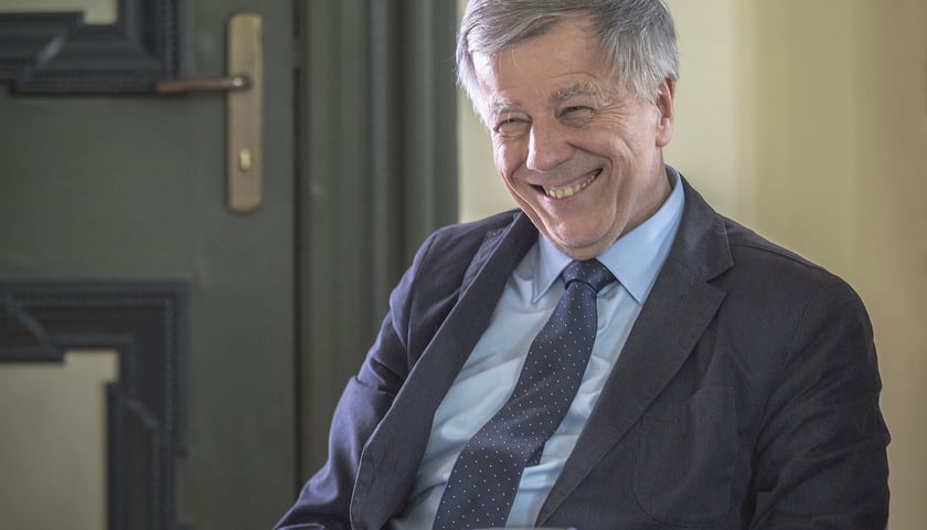 Uśmiechnięty prof. Jan Miodek siedzi przy biurku, ubrany w ciemny garnitur, jasną koszulę i krawat w kropki