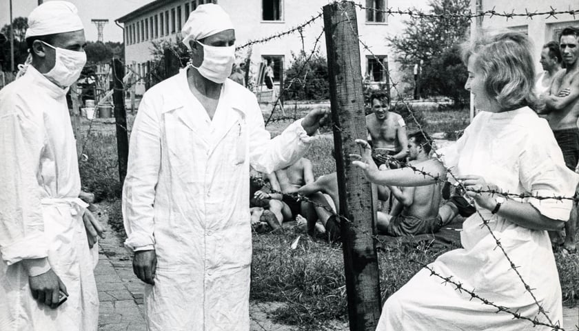 Zdjęcie z wystawy „Odizolowani 1963. Epidemia ospy we Wrocławiu” w Pałacu Królewskim. Widać dwóch lekarzy w białych fartuchach i maseczkach oraz lekarkę w białym uniformie. Stoją przy płocie z drutem kolczastym.