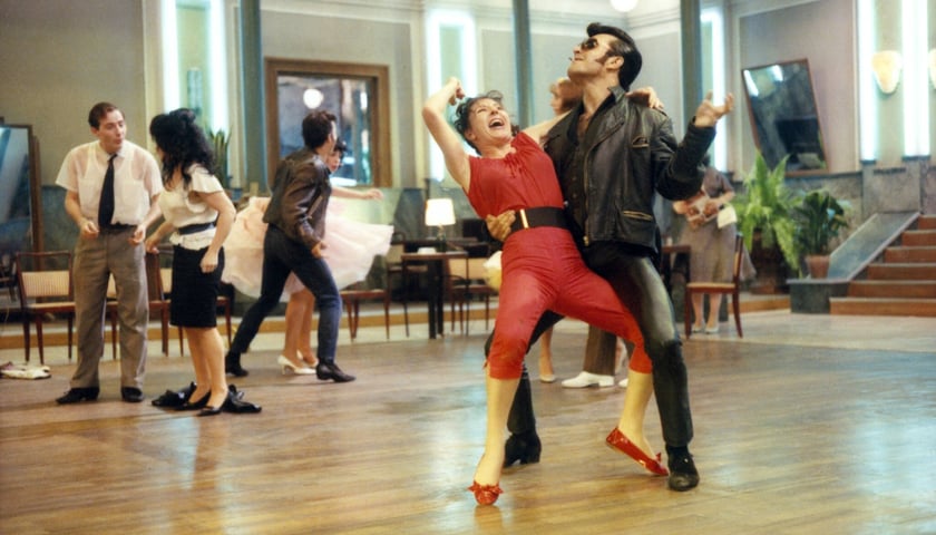 Mężczyzna w czerni stylizowany na Elvisa Presleya i kobieta w czerwonym kombinezonie tańczą na parkiecie w filmie „Bal”