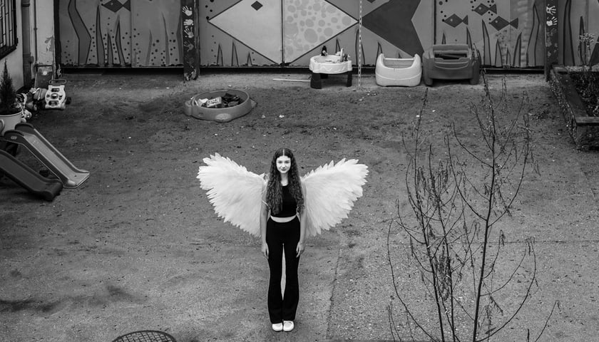 Młoda kobieta z doczepionymi anielskimi skrzydłami stoi pośrodku pustego podwórka. Zdjęcie czarno-białe