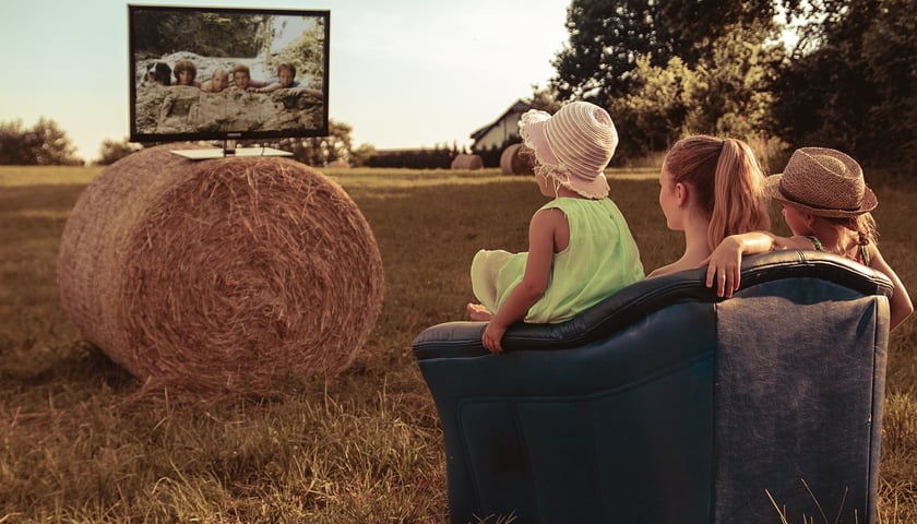 Trójka dzieci siedzących na fotelu i oglądających film. Telewizor ustawiony jest w plenerze, na beli siana. Zdjęcie ilustracyjne 