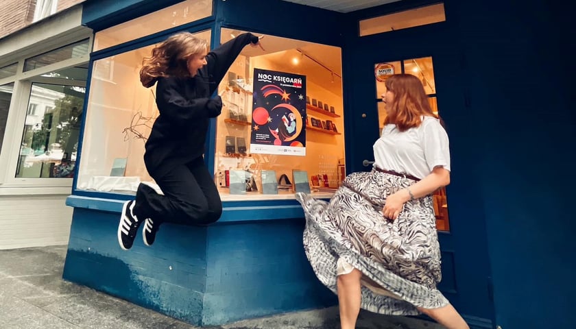 Dwie młode kobiety tańczące przed księgarnią, w której witrynie plakat Nocy Księgarń 2023 