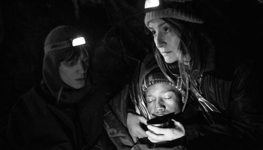 Powiększ obraz: Kadr z filmu "Zielona granica". Julia (Maja Ostaszewska) pomaga nocą w lesie uchodźcom. Jednemu podtrzymuje głowę. Z lewej aktywista