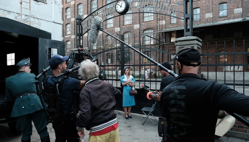 Kobieta ubrana w niebieską sukienkę w stylu lat 30. trzyma na rękach niemowlę i stoi przed bramą, przed nią ekipa filmowa 